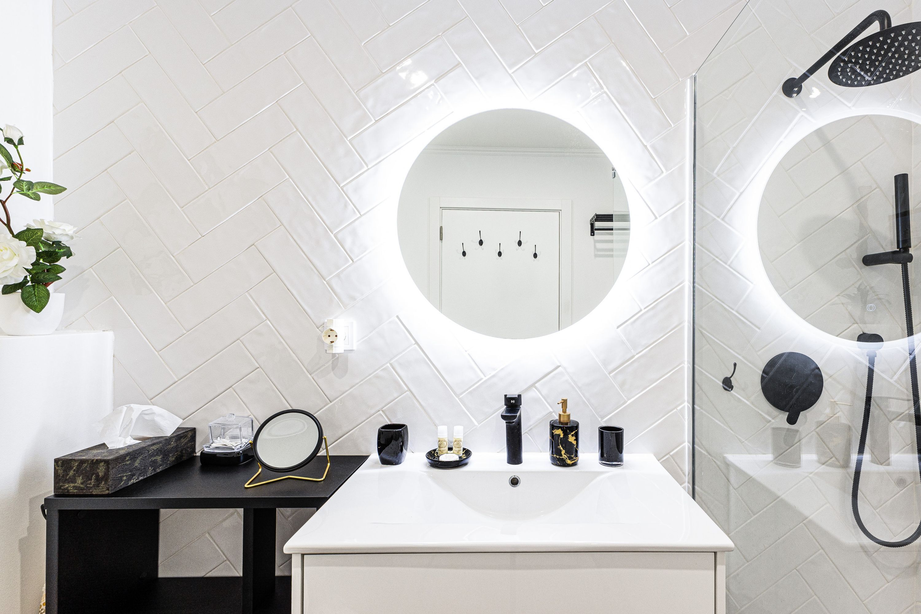 A modern bathroom with a white sink, a round mirror, a handheld showerhead, and a toilet visible through a glass partition.
