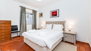 Ein Schlafzimmer mit einem großen Bett, zwei Nachttischen, einer Holzkommode, einem Sessel und einem Fenster mit Vorhängen.