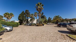 A paved parking area with several cars, palm trees, and a building in the background. - Porto de Mos - Nucleo 2 (Lagos)