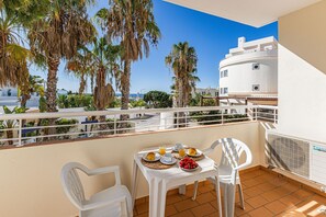 Ein Balkon mit einem Frühstückstisch, der auf eine Küstenlandschaft mit Palmen und einem weißen Gebäude blickt.