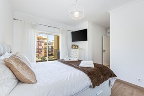 Ein Schlafzimmer mit Bett, Flachbildfernseher, Kommode, einem Fenster mit Vorhängen und Balkonblick.