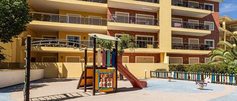 A multi-story building with balconies, a playground with a slide, and a pool area.