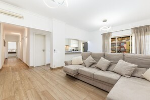 Ein modernes Wohnzimmer mit einem beigefarbenen Ecksofa, Holzboden, einer Küche im Hintergrund und einem großen Fenster mit Vorhängen.