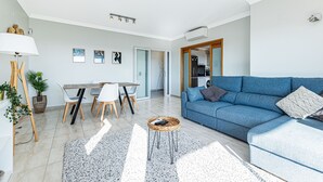 Ein modernes Wohnzimmer mit einem blauen Sofa, einem Esstisch mit Stühlen, einem Couchtisch und einem durch eine offene Tür einsehbaren Küchenbereich.