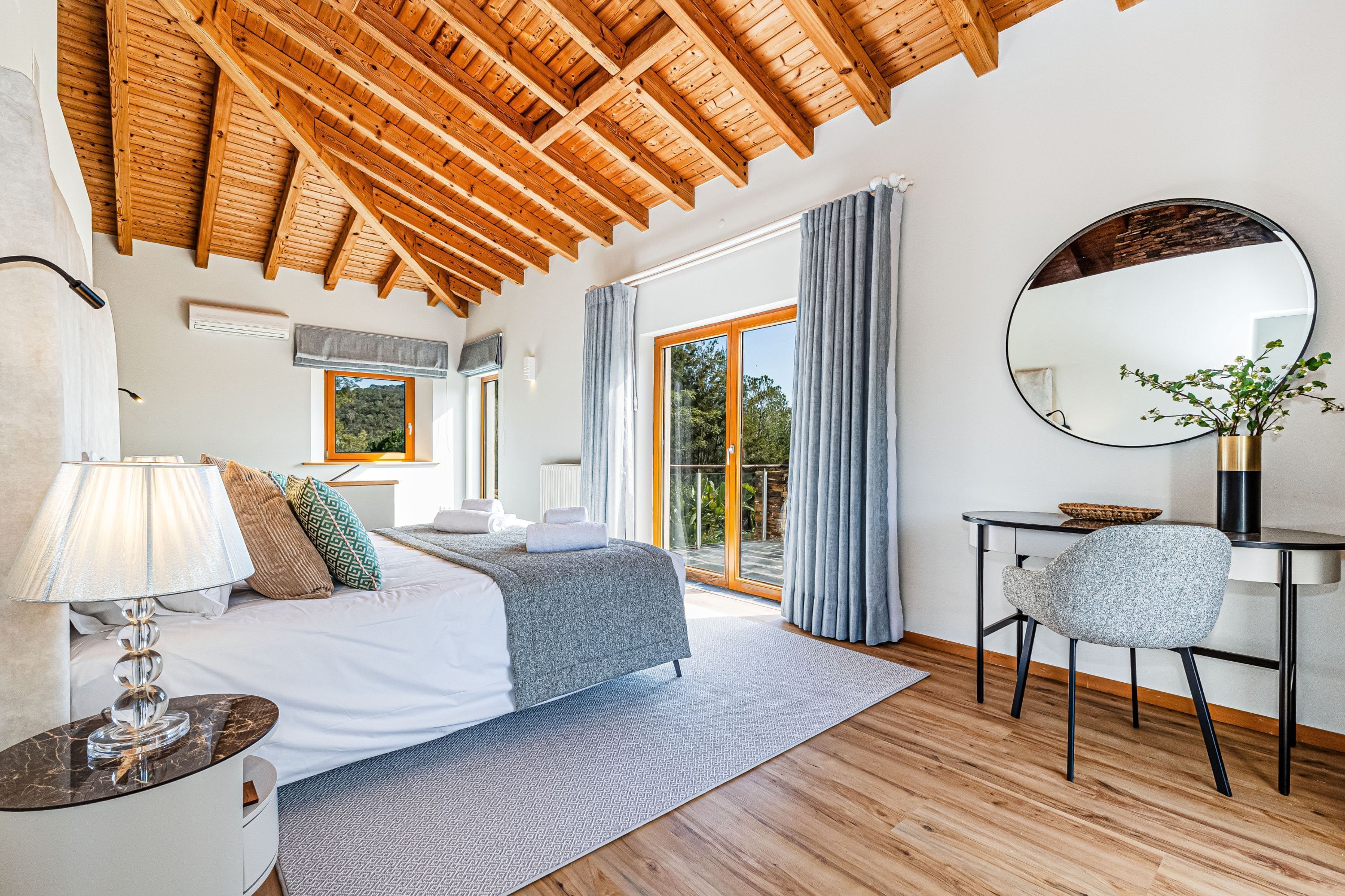 Une chambre avec un plafond en bois, un lit recouvert d’une couverture grise, un bureau avec une chaise et une grande fenêtre avec des rideaux.