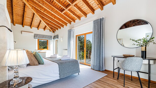A bedroom with a wooden ceiling, a bed with a grey blanket, a desk with a chair, and a large window with curtains. - Casa Corsino in Marmelete (Monchique)