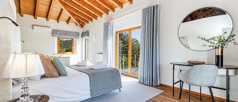 Une chambre avec un plafond en bois, un lit recouvert d’une couverture grise, un bureau avec une chaise et une grande fenêtre avec des rideaux.