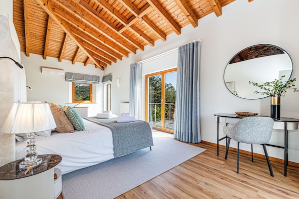 A bedroom with a wooden ceiling, a bed with a grey blanket, a desk with a chair, and a large window with curtains. - Casa Corsino in Marmelete (Monchique)