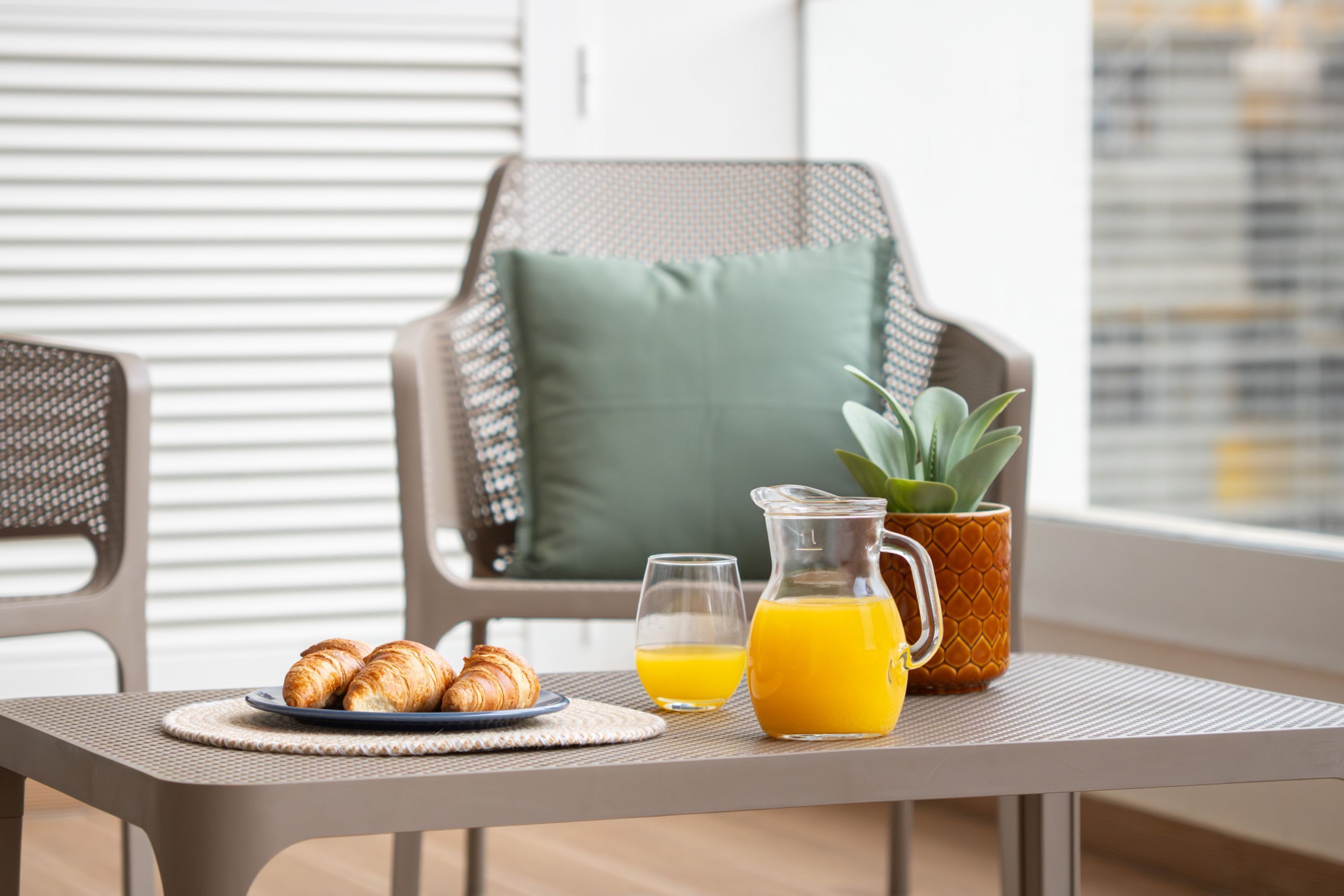 Desayuno con croissants, jugo de naranja y una jarrón sobre una mesa, con una silla y una planta al fondo.