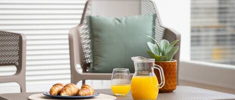 Desayuno con croissants, jugo de naranja y una jarrón sobre una mesa, con una silla y una planta al fondo.