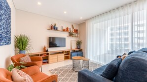 A modern living room with a TV, orange and blue sofas, a coffee table, and a large window with sheer curtains. - Adega Luxury Near Lagos City Center With Pool (Lagos)