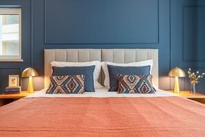 Un letto con una testiera beige, due cuscini a motivi geometrici, una coperta color pesca e due abat-jour.