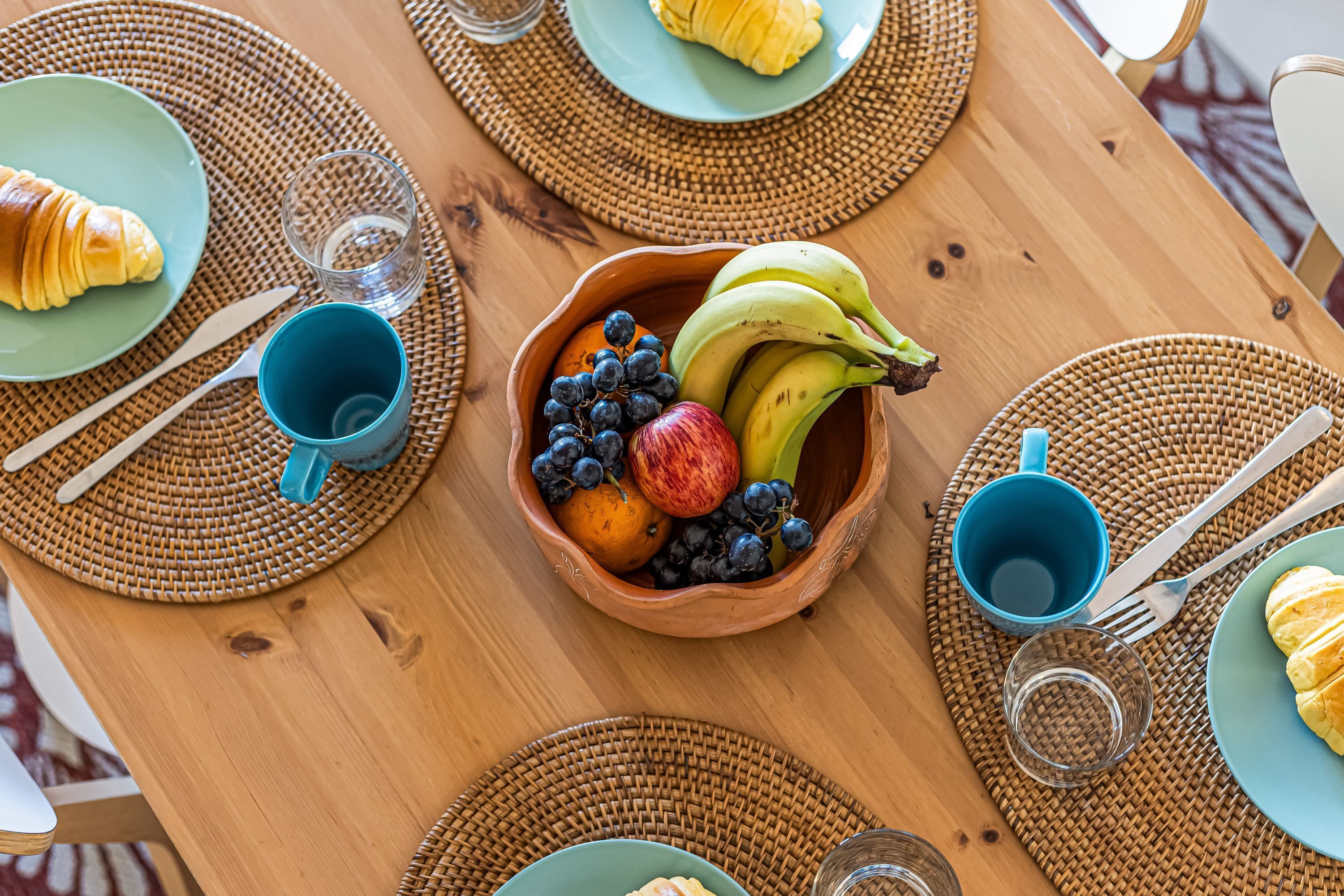 A wooden table with a woven placemat, a bowl of fruit, a croissant, and a blue cup.