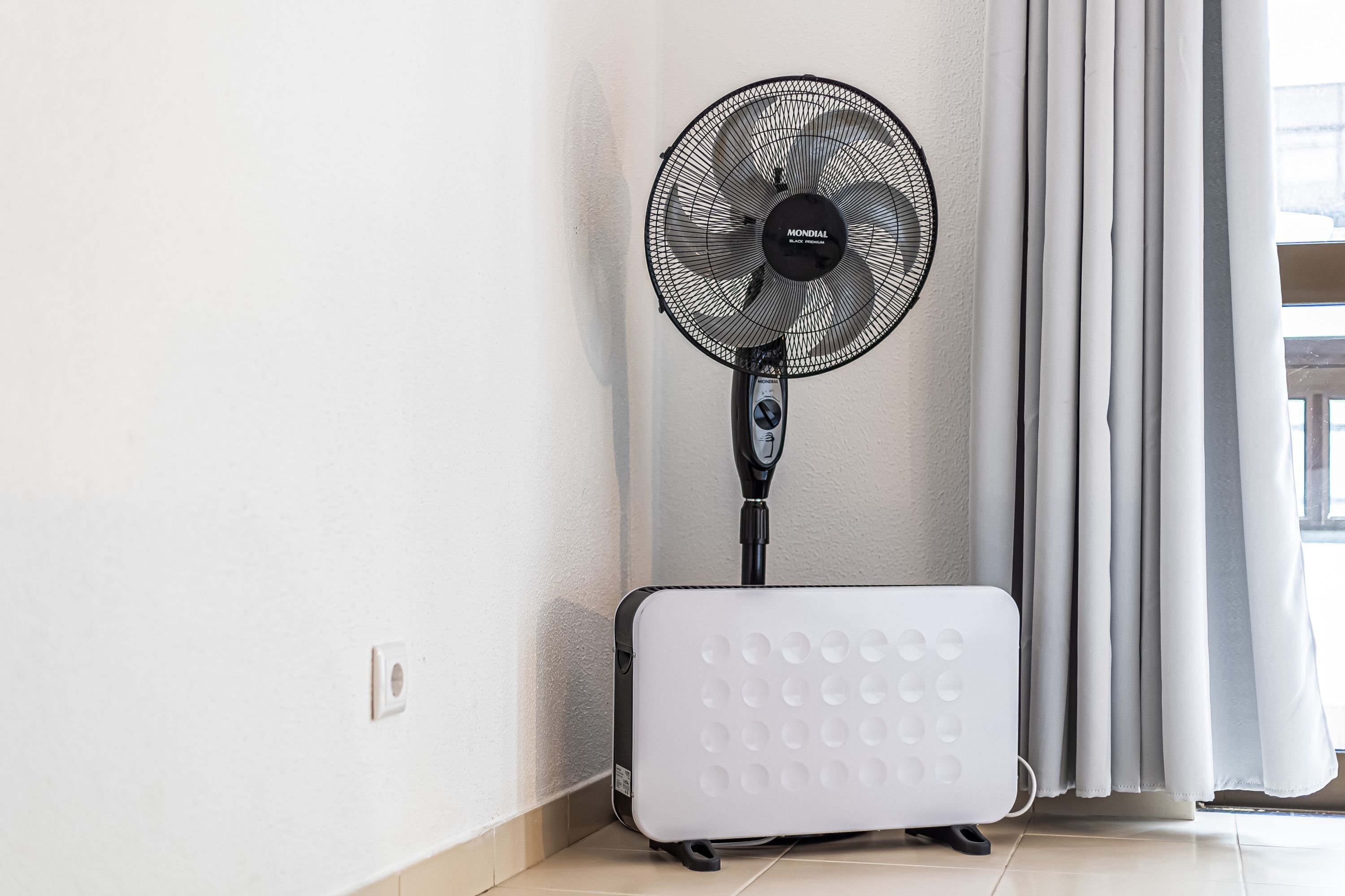 A standing fan, a white portable heater, and a window with curtains.