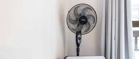 A standing fan, a white portable heater, and a window with curtains.