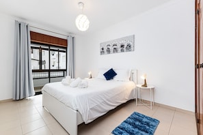 Ein modernes Schlafzimmer mit einem großen Bett, einem Nachttisch mit Lampe, einem Gemälde an der Wand und Blick durch ein Fenster.