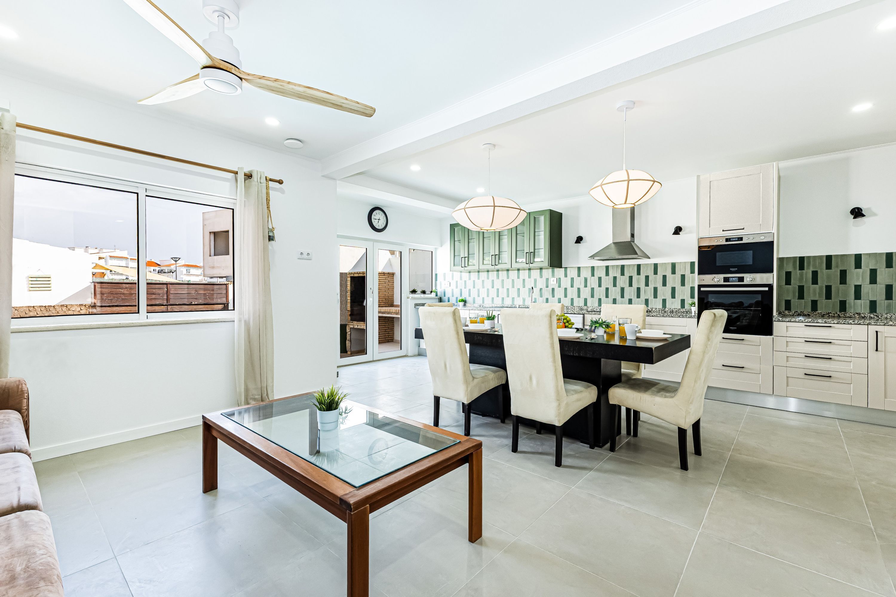 Una cocina moderna con armarios blancos, una zona de comedor con mesa de cristal y un ventilador de techo.