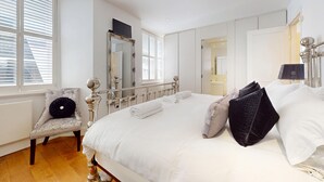 A bedroom with a large bed, a chair, a mirror, and a window with blinds. - Central 2BR Home Steps to Kensington Palace & Hyde park (London)