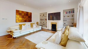 A modern living room with a white sofa, a fireplace, a flat-screen TV, and bookshelves. - Central 2BR Home Steps to Kensington Palace & Hyde park (London)