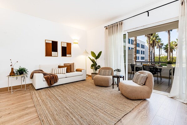 Ein modernes Wohnzimmer mit einem weißen Sofa, einem beigefarbenen Sessel, einem großen Teppich und einem Essbereich, der durch eine Glastür sichtbar ist.