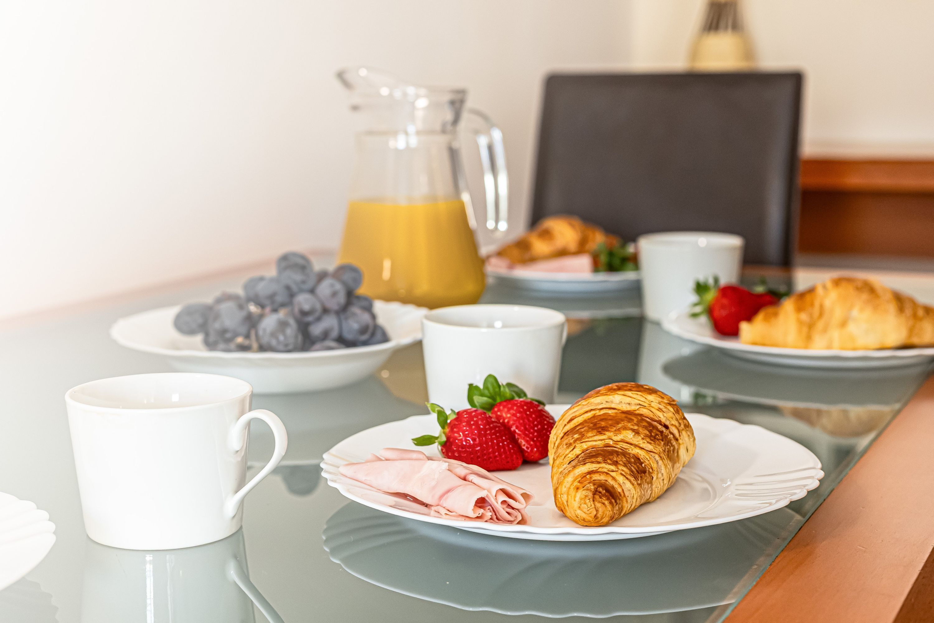 Une table mise pour le petit-déjeuner, avec une table en verre, des assiettes remplies de nourriture, un pichet de jus d’orange et une tasse.