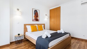 A neatly made bed with a wooden headboard, a small round side table with a plant, and a painting on the wall. - Encosta do Vau in Portim o (Portimão)