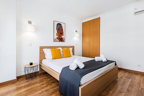A neatly made bed with a wooden headboard, a small round side table with a plant, and a painting on the wall. - Encosta do Vau in Portim o (Portimão)