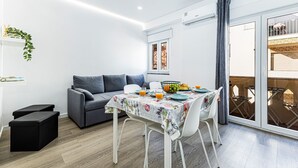 Una sala de estar moderna con un sofá gris, una mesa de comedor con mantel floreado y un balcón con mobiliario de madera.