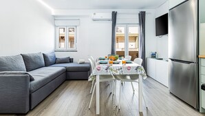 Una sala de estar moderna con un sofá gris, una mesa de comedor con sillas, una zona de cocina con refrigerador y un televisor montado en la pared.
