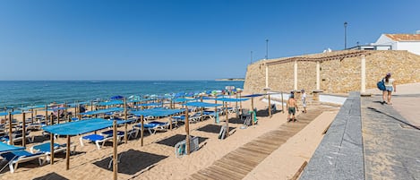 A beach with blue umbrellas and sun loungers, a stone wall, and a wooden walkway.