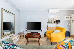 Ein Wohnzimmer mit einem großen Flachbildfernseher, einem gelben Sessel, einem Couchtisch aus Holz und einem Sofa mit floralen Kissen.
