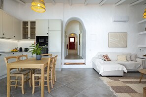 A modern kitchen with white cabinets, a dining table with chairs, a potted plant, and a hallway leading to a living area with a sofa and a rug. - Casa Oliva Ref gio Rural Contempor neo (Beja)