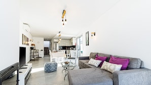 A modern living room with a grey sofa, ottoman, and a flat-screen TV. There is a dining area with a table and chairs, and a kitchen in the background. - Casa Colina II Pool BBQ Family Retreat (Silves)