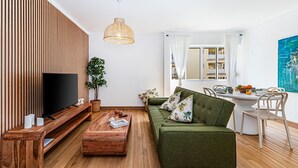 Moderni olohuone, jossa on litteänäyttöinen televisio, puupaneloitu seinä, vihreä sohva, ruokapöytä ja suuri ikkuna, jossa on verhot.