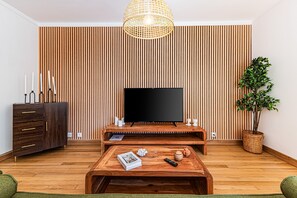 A modern living room with a flat-screen TV, wooden entertainment unit, a coffee table with books and a remote, a green sofa, and a potted plant. - Casa Sol Vilamoura (Loulé)