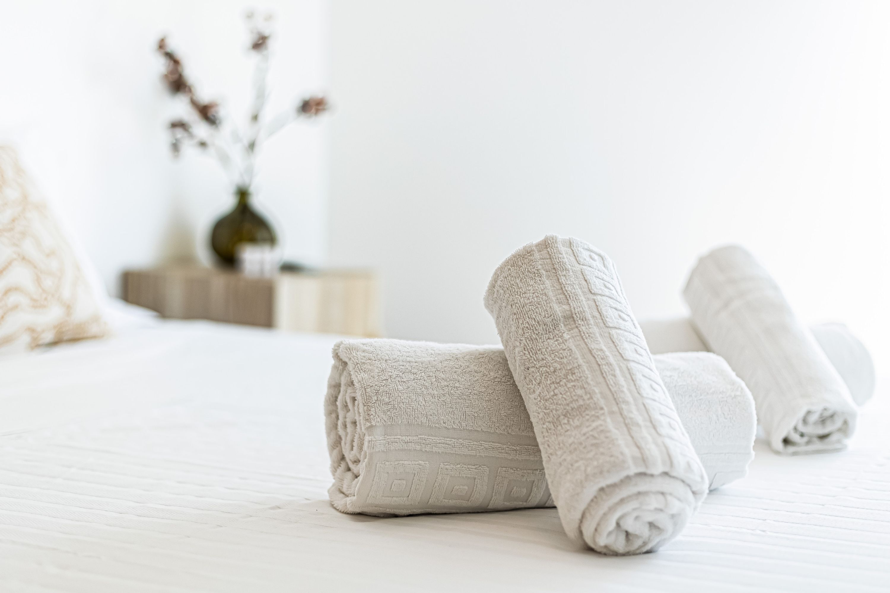 A neatly made bed with rolled white towels and a vase with dried flowers in the background.