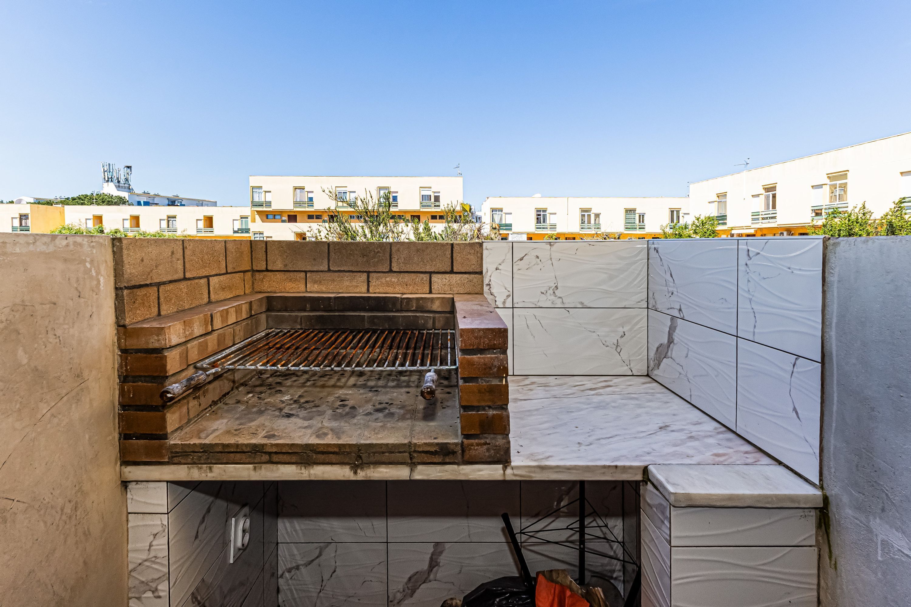 Een dakbarbecueplaats met een bakstenen grill, betegelde muren en uitzicht op gebouwen op de achtergrond.