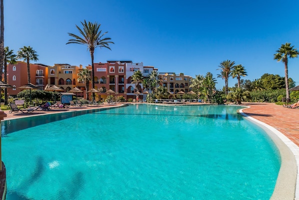 A swimming pool with lounge chairs and umbrellas, surrounded by palm trees and a hotel building with multiple floors and balconies. - Jardins da Meia Praia Villa Pool Family (Lagos)
