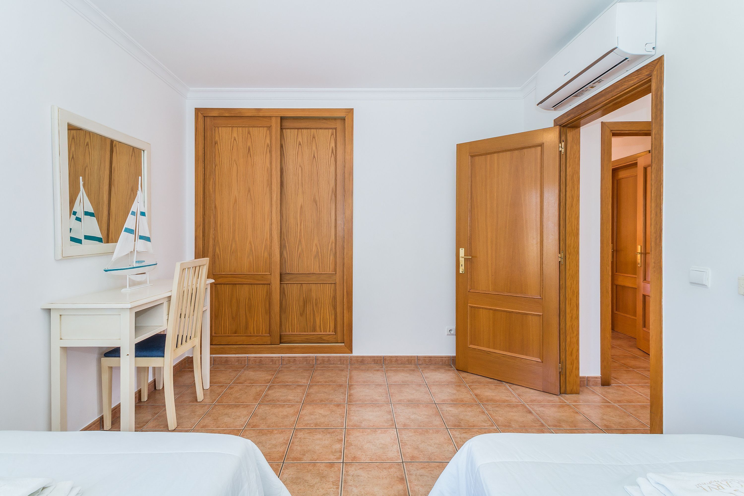 A room with a desk, chair, two beds, wooden wardrobe, and an air conditioning unit.