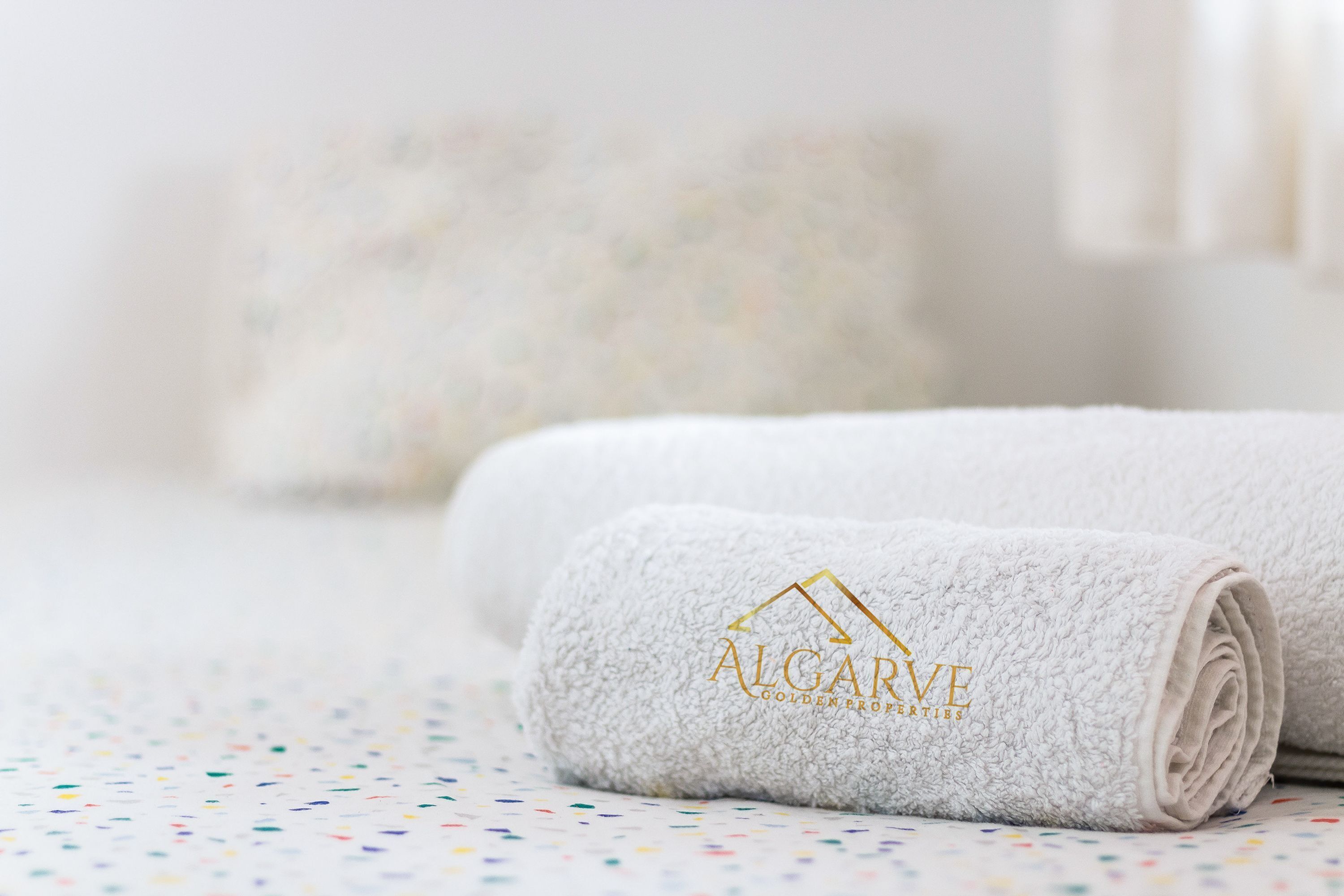 A neatly folded white towel with the logo 'ALGARVE GOLDEN PROPERTIES' on a bed with a colorful polka dot bedspread.