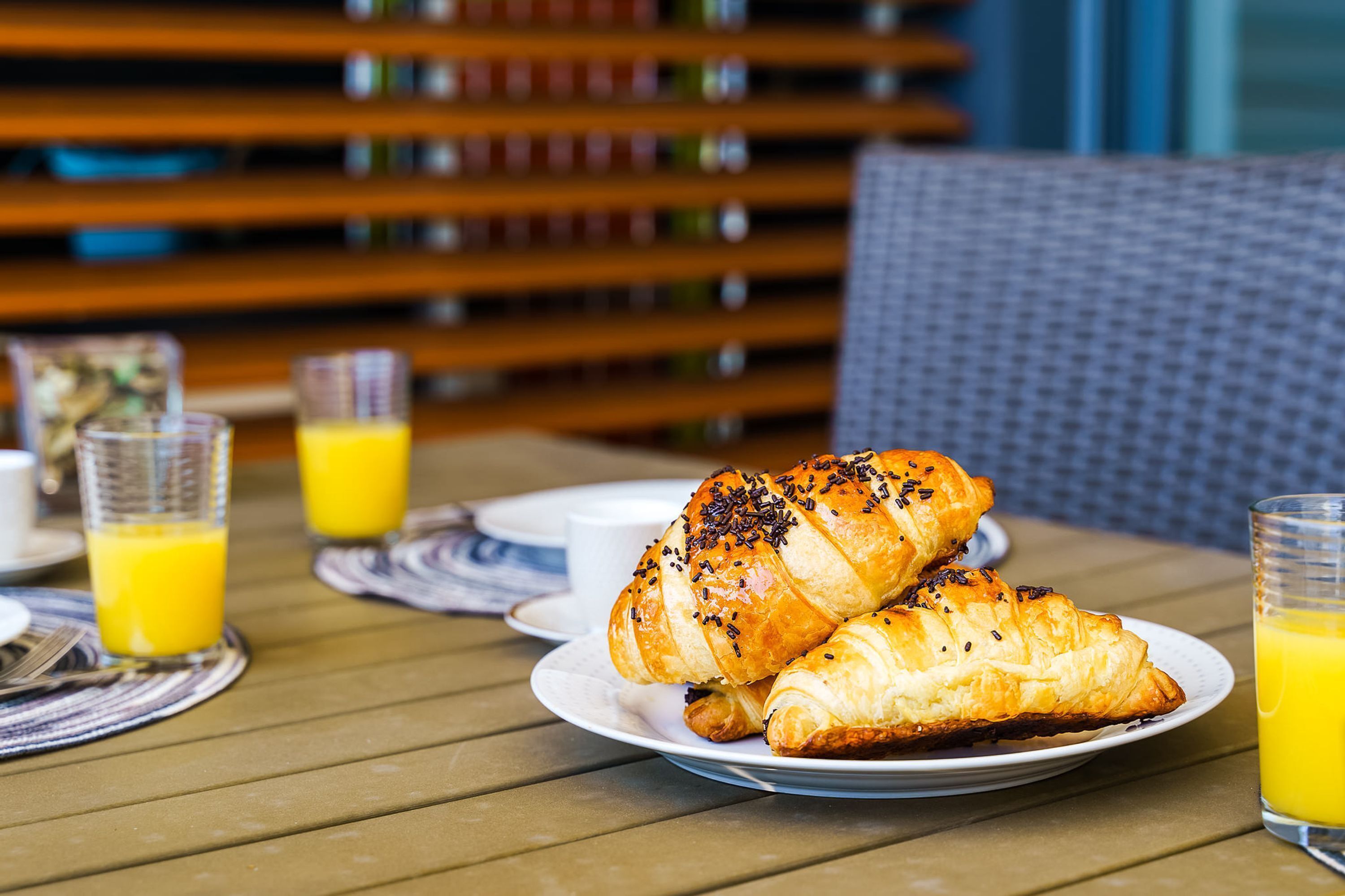 A breakfast setting with croissants on a plate, a glass of orange juice, and a wicker chair in the background.