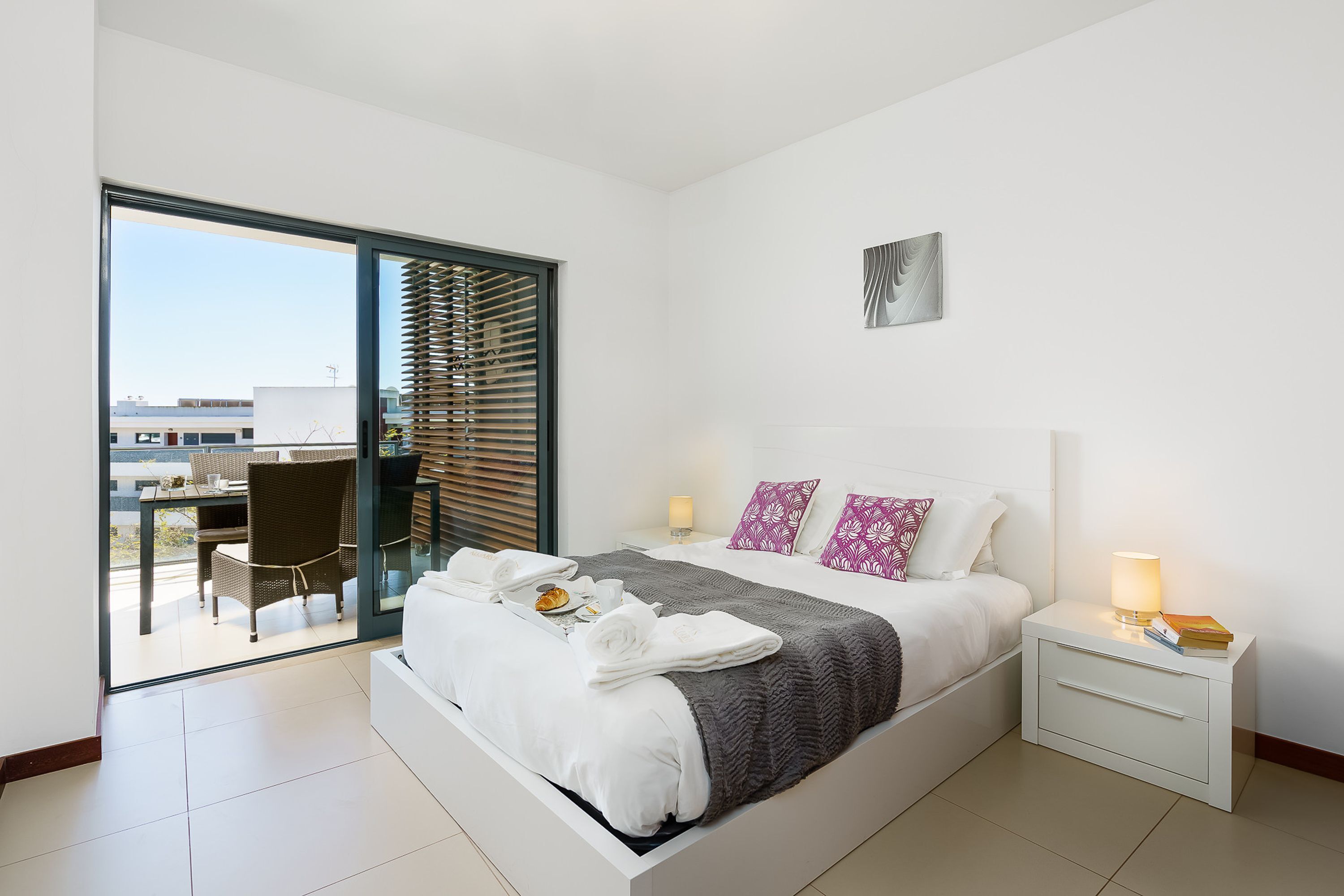 A modern hotel room with a large bed, a bedside table with a lamp, a view of the balcony, and a dining area.