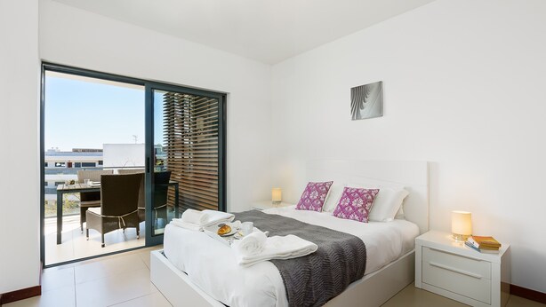 A modern hotel room with a large bed, a bedside table with a lamp, a view of the balcony, and a dining area. - Marina Village Pool Garage Prime Location AC (Lagos)