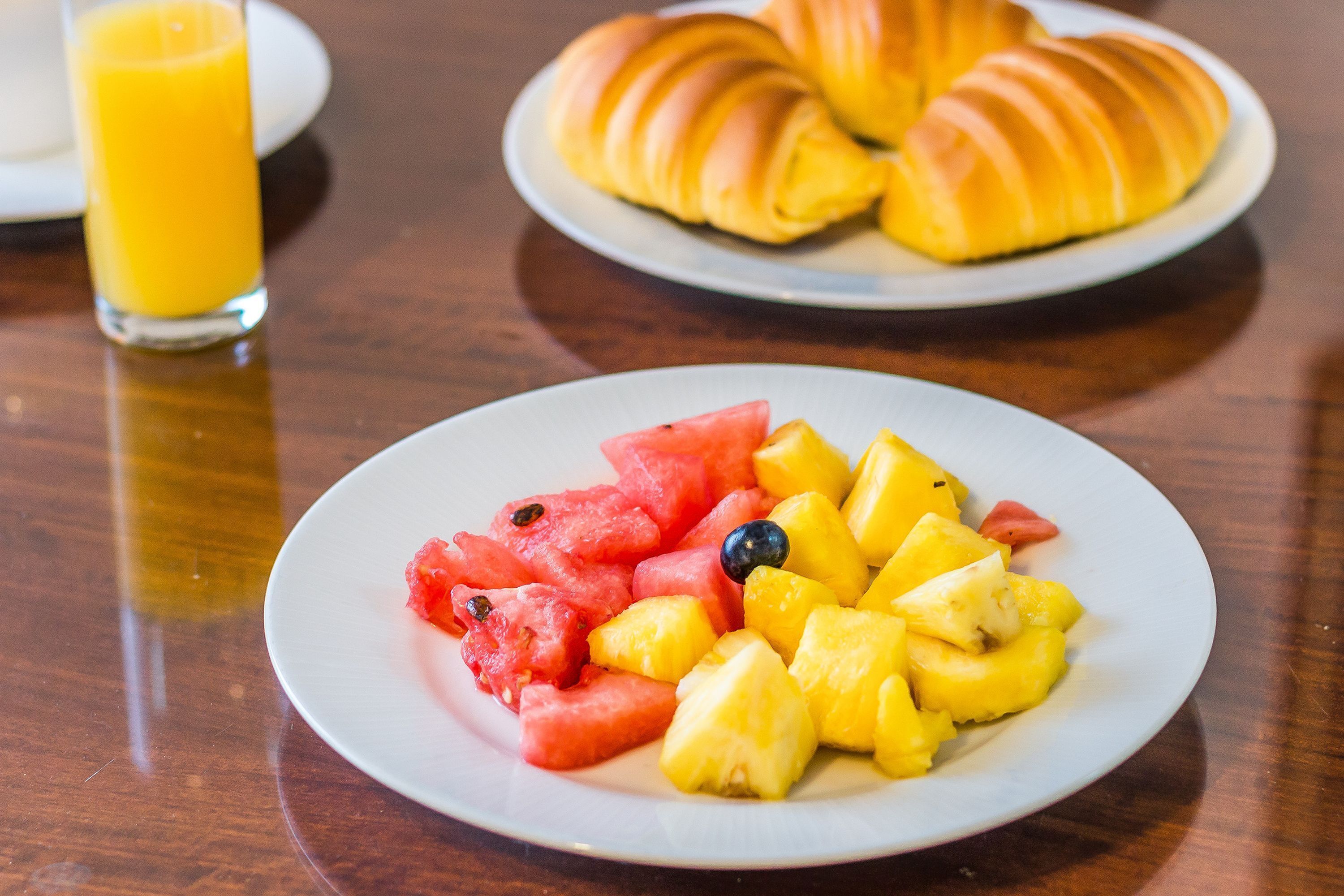 Un piatto con anguria, ananas e mirtilli, un bicchiere di succo d'arancia e croissant su un tavolo di legno.