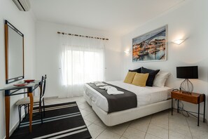 Um quarto com cama, mesa de cabeceira, luminária, escrivaninha, cadeira e um quadro na parede.