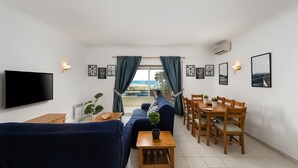 Uma sala de estar com sofá, TV, mesa de jantar, cadeiras e vista para o exterior.