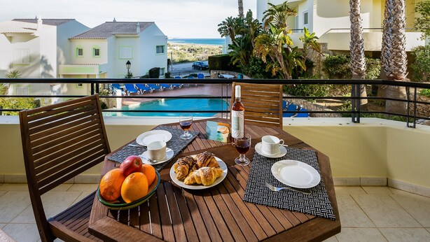 Uma mesa de jantar com itens de café da manhã, uma garrafa e vista para uma piscina e edifícios.