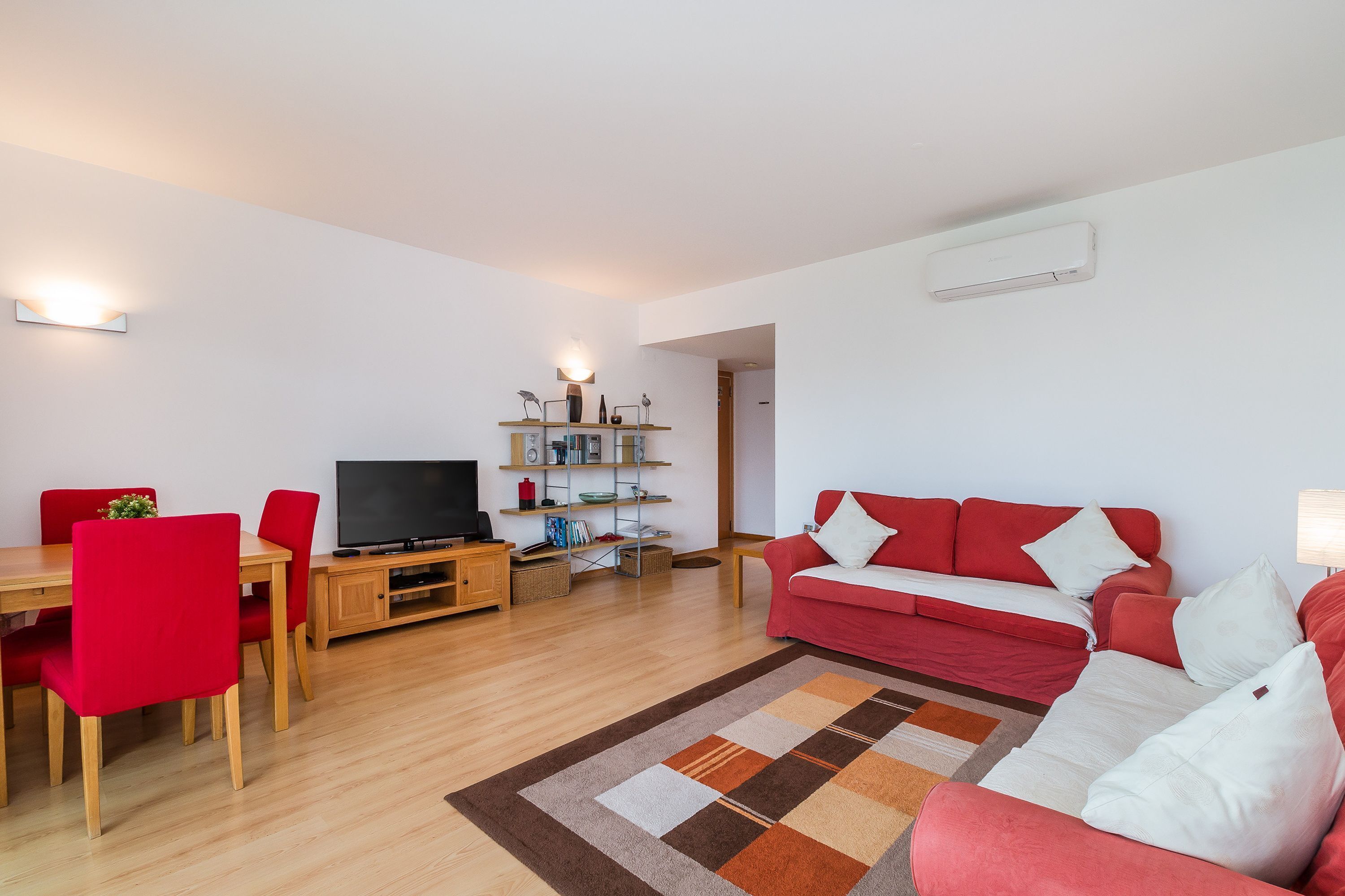 Una sala de estar con un sofá rojo, un televisor, una mesa de comedor de madera y sillas rojas, y una estantería.