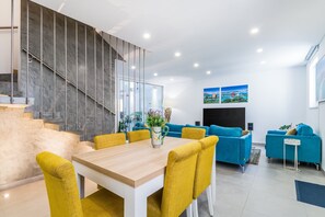 A modern living room with a wooden dining table, yellow chairs, blue sofas, and a staircase with a glass railing. - Vista Bonita Dowtown Family (Lagos)