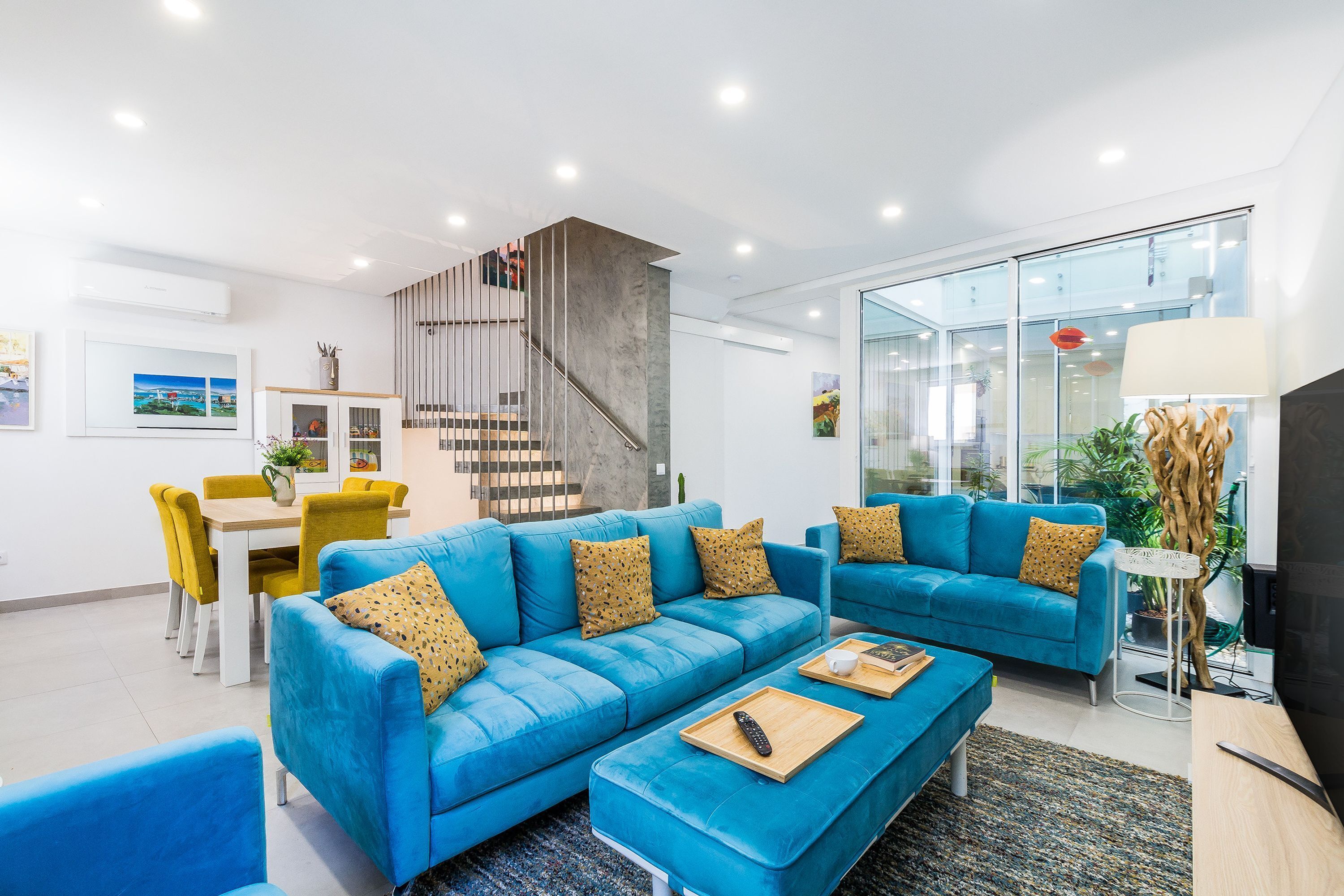 A modern living room with a blue sofa, a dining table with yellow chairs, a staircase, and a large window.
